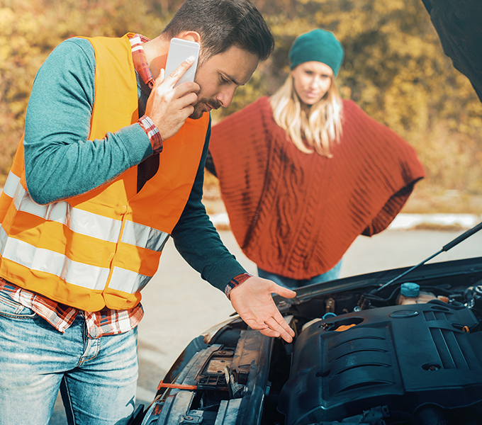 a recovery man looking at the engine of a car with a woman who owns the car in the background
